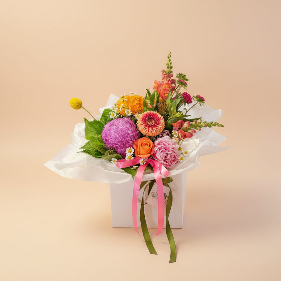 Bright colourful flowers arranged in a white bloom bag with pink and green ribbons and tissue paper with a soft peach background.